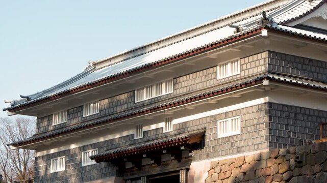 Low angle tilt up of traditional Kahoku-mon gate at historic Kanazawa castle park with stone wall and tile roof in Japan.