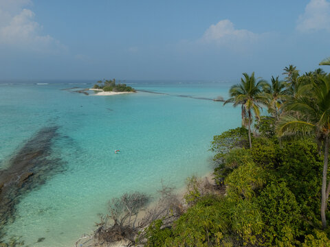 Aerial view of pristine turquoise waters gently lapping a palm-fringed shore, with a distant sandy islet connected by a shallow reef, Thuraakunu, Thiladhunmathee Uthuruburi, Maldives.