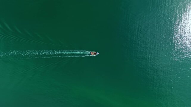 A small boat moves across calm water under clear blue skies. Waves trail behind as it glides smoothly. Sunlight reflects off the surface, creating bright patches of light.