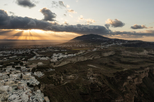 Aerial view of sunlight piercing through dark clouds over the whitewashed buildings of Santorini and the rugged landscape, Thira, Greece.