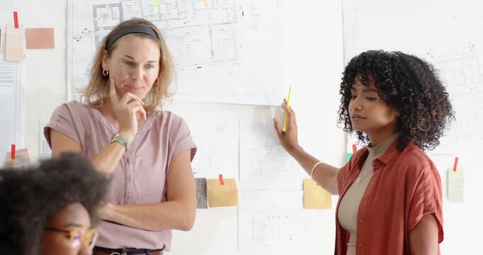 Diverse female team viewing pinned plans as curly woman pointing yellow pencil seated peer drawing