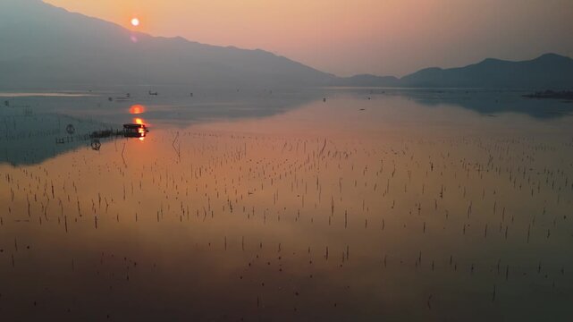 Golden hour sunset illuminates a calm lake, creating an orange reflection across the water, with countless aquaculture poles and a floating house dotting the tranquil expanse under hazy mountains