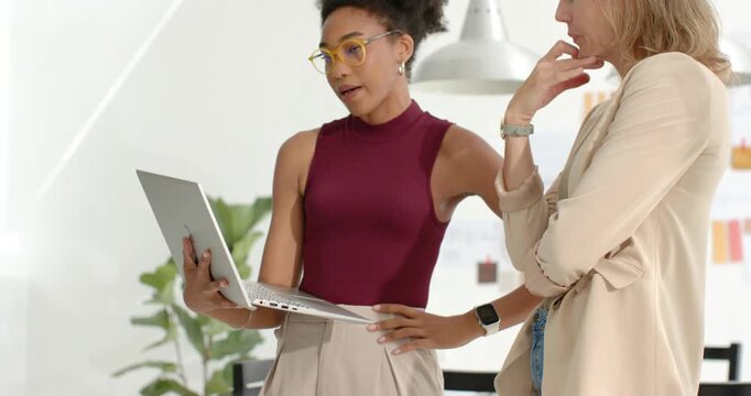 Two female coworkers in smart clothes in office, laptop-holder adjusting silver laptop for review