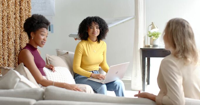 Friends on sofa, center woman in yellow after blazer gesturing, alternating typing on laptop noting