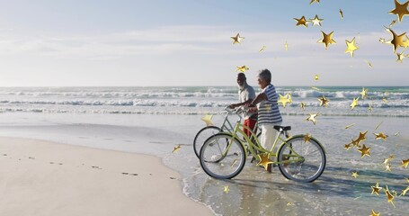 Fototapeta premium Walking couple wearing striped top holding cruiser bikes at surf with gold stars, copy space