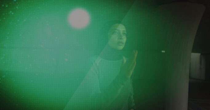 Touching woman in dark headscarf facing green pixel-grid panel by curved concrete wall under light
