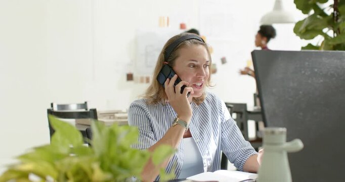 Adult woman answering mobile call while referencing monitor, papers and keyboard during work call