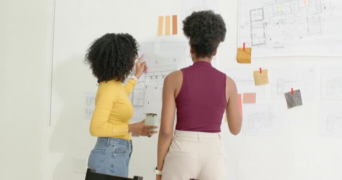 Diverse female designers reviewing plans at pin-up wall, pointing with pencil, moving swatches