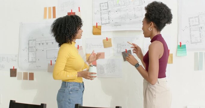 Diverse female designers seeing pinned plans by wall pointing swapping swatch refining layout