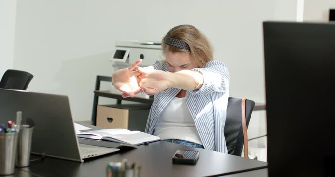 Mid-30s woman in striped shirt stretching arms after long laptop work at office desk with notebook