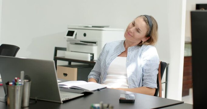 Mid-adult woman leaning back, raising arms and stretching wrists at desk with laptop easing tension