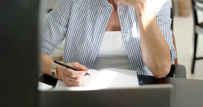 Middle-aged woman taking notes while reviewing laptop to finish work at desk, holding notebook pen