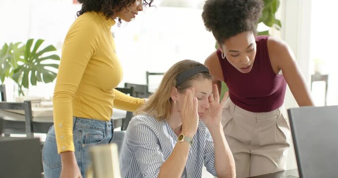 Diverse female team coming on seeing coworker rubbing temples at desk, pointing to monitor to help