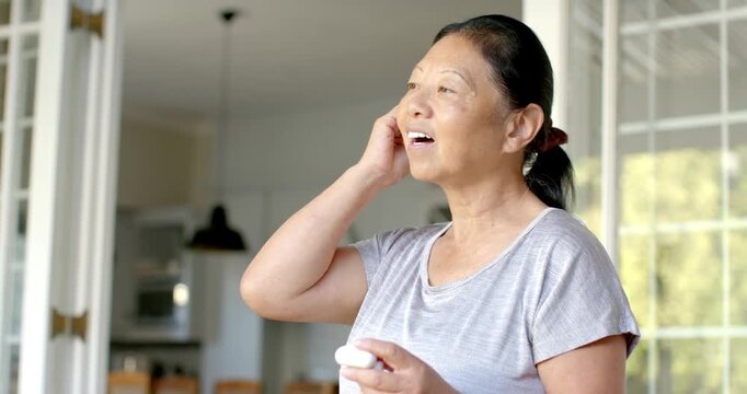 Senior Asian woman hearing chime, holding case, inserting earbuds, confirming fit at home doorway