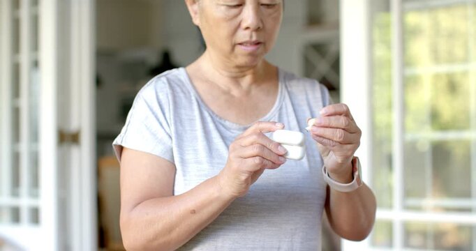 Senior Asian woman at home opening charging case, removing earbud, inserting and adjusting for call
