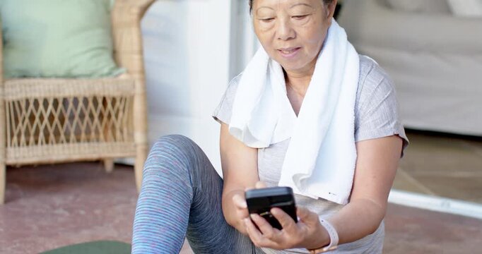 Senior Asian woman tapping smartphone after short workout, smiling on patio mat in workout gear