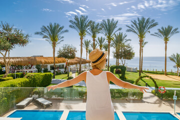 Obraz premium Back view of a girl standing on a terrace of a luxury tropical resort, enjoying the ocean view and sunny weather during her summer vacation trip