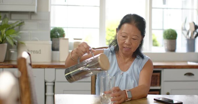 Senior Asian woman reaching for pitcher, pouring water into glass at sunlit kitchen table to drink