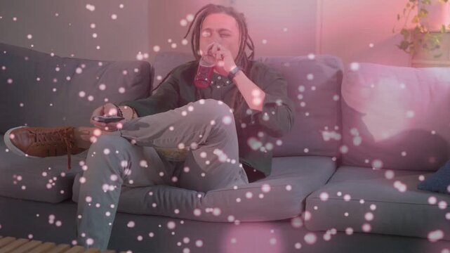 Vertical video: Man pressing remote, surfing TV, sipping bottle on couch, pink bokeh veiling scene