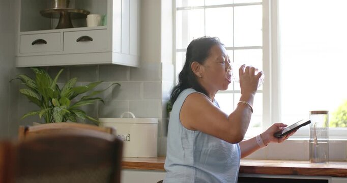 Senior woman standing at kitchen counter holding tablet and glass, sipping and reacting to message