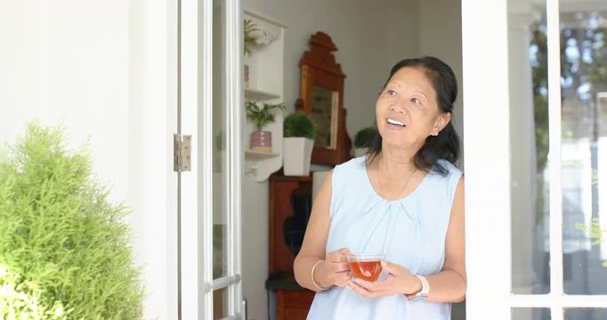 Senior woman standing in doorway, sunlight drawing gaze outward, lifting glass cup and sipping tea