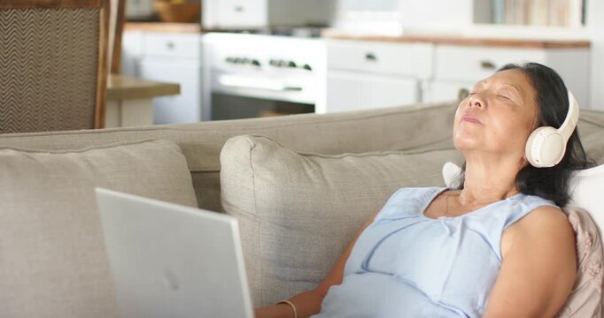 Hearing laptop audio Senior Asian woman with headphones closing eyes to rest on sofa, copy space
