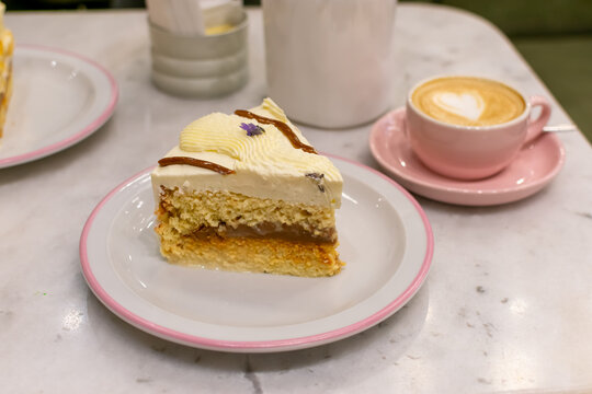 Porci&oacute;n de torta tres leches y vainilla con relleno de dulce de leche y glaseado blanco. Interior minimalista de cafeter&iacute;a con caf&eacute; decorado con forma de coraz&oacute;n. Imagen con desenfoque.