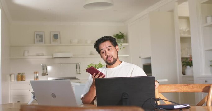 African American man reading red smartphone alert, responding and checking laptop at kitchen table