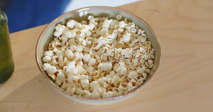 Mid-adult African American man reaching into bowl, scooping popcorn, snacking at table with bottle