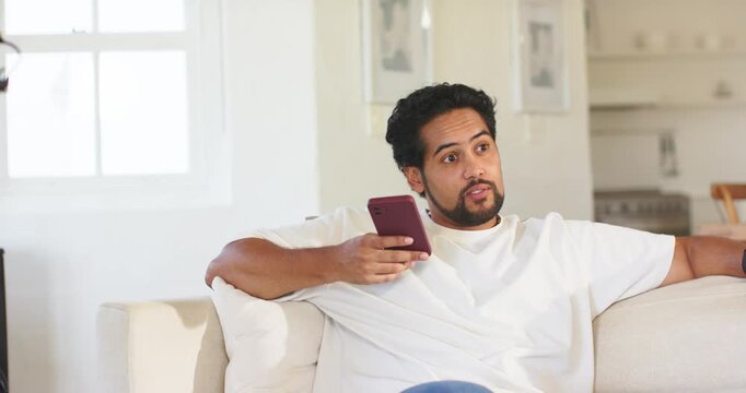 Mid-adult African American man sitting on home sofa holding maroon phone and reacting to alert