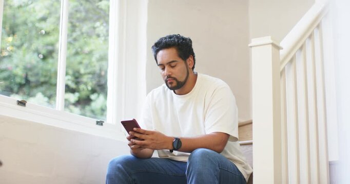 African American man starting looking at phone, sitting on stairwell by window, smiling at message