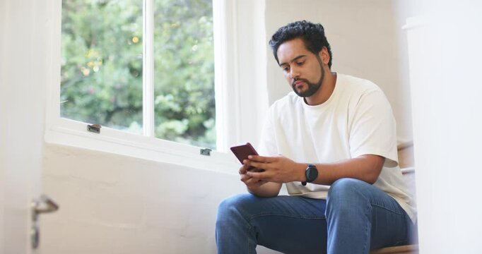 African American mid adult man sitting on stair landing by window, tapping reddish-cased smartphone