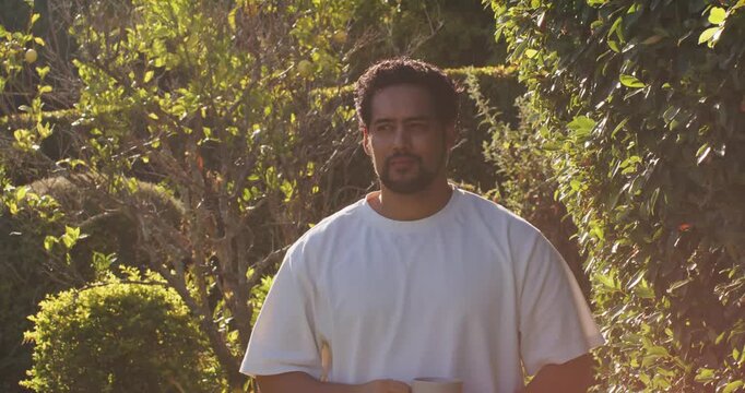 African American man white T holding mug in garden turning head to off-camera cue sun flaring