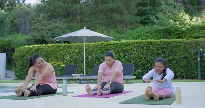 Diverse female family with girl following center adult's cues, stretching on patio on yoga mats