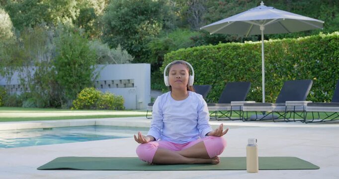 African American child hearing audio in headphones, meditating on mat by pool, camera tightening