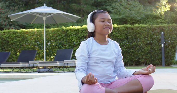 African American girl child meditating on mat beside pool, headphones playing guided breathing