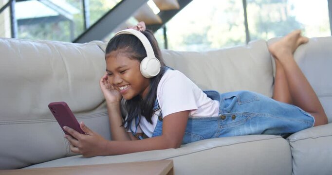 Receiving funny audio, youth female lying on sofa holding smartphone, laughing, touching headphones