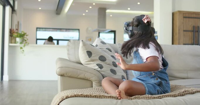 African American mom watching as youth girl in overalls reaching following VR headset cues on sofa
