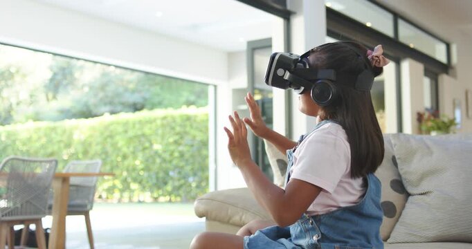 Diverse mom and daughter, girl responding to VR reaching items above sofa while mom with mugs