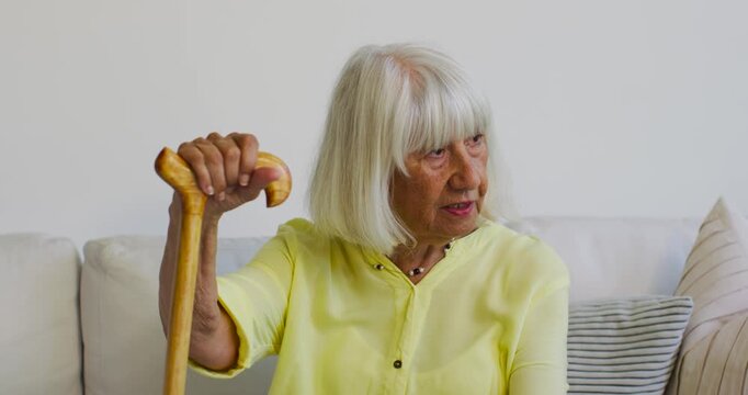 Senior female wearing blouse holding wooden cane on sofa, lowering gaze and turning right, thinking