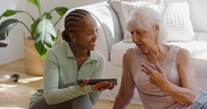 Diverse female carer and senior woman sitting at home showing tablet while helping and talking
