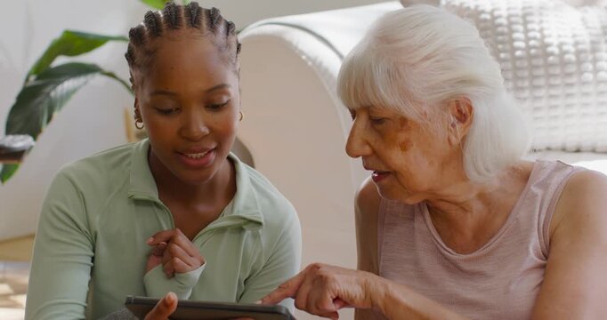 Diverse female aide showing tablet on sofa while senior woman pointing at screen, learning use