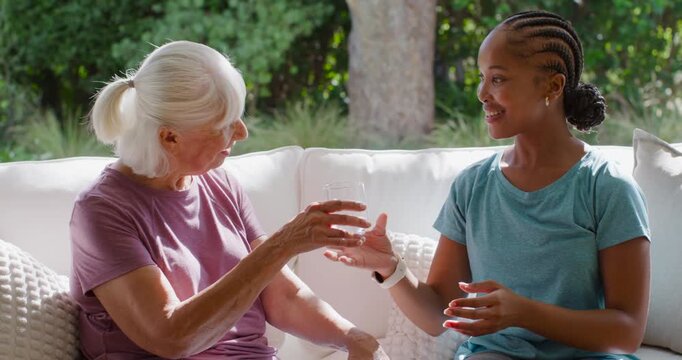 African American caregiver handing clear glass to senior woman on patio sofa, hydrating senior