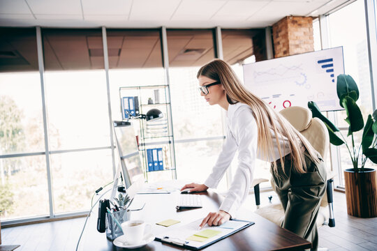 Young professional woman leaning over desk in modern office reviewing data on computer and planning strategy