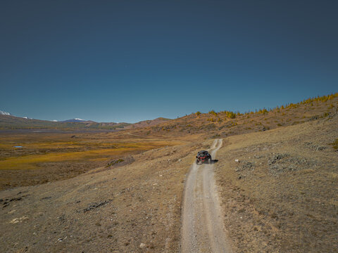 Off-road buggy UTV vehicle driving on a winding dirt road through a vast mountain valley in autumn. Aerial view of extreme adventure trip in wild remote highlands under a clear blue sky.