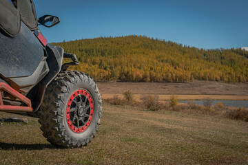 Obraz premium Close-up of off-road buggy UTV wheel with red beadlock parked on a field against a background of autumn larch forest and lake. Extreme adventure trip concept in wild nature under blue sky.
