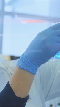 Vertical video: Gloved arm entering from left, lifting blue vial toward window, inspecting sample