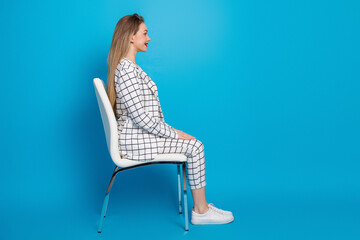 Businesswoman sitting upright in profile on a white chair against a bright blue background wearing a checked suit and white sneakers © deagreez