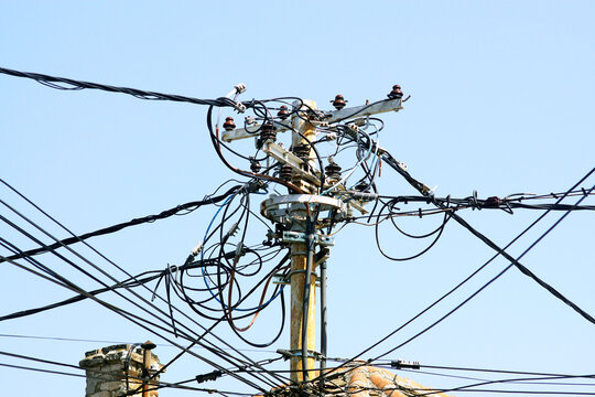 Rusted utility pole crowded with looping cables and ceramic insulators against sky. Captures failing grid infrastructure, electrical distribution chaos, and urgent power network modernization needs.
