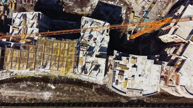 Cranes work over a construction site where high-rise buildings rise. Workers handle materials on the ground and the area is busy with ongoing construction.